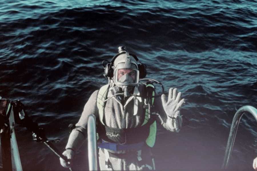 A deep-sea diver in a full-body protective suit and helmet waves at the camera while climbing up a metal ladder from dark ocean waters. The diver is wearing a rebreather system, gloves, and a safety harness marked “Sea Quest.”