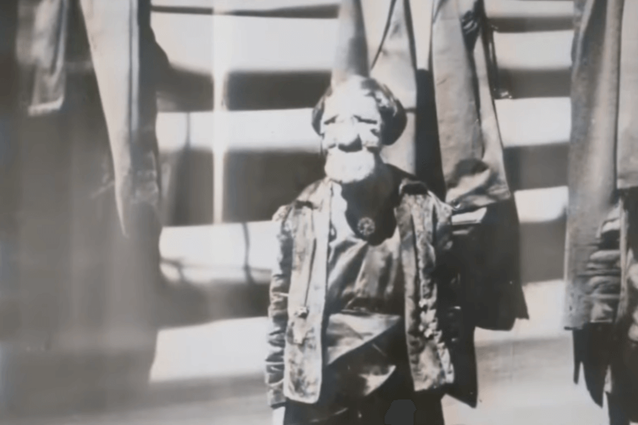 Vintage black and white photo of a person with dwarfism and a visibly disfigured face, standing in front of a staircase flanked by coats or garments hanging on either side. The person wears formal or performance-like attire, possibly suggesting involvement in public exhibition or a sideshow, reflecting a time when individuals with visible differences were often objectified for entertainment.
