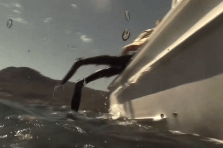 Low-angle shot from the water showing a diver in fins being pulled or lifted onto the side of a boat, with only their legs visible above the surface. Water droplets on the lens and a mountainous coastline in the background add to the action feel.