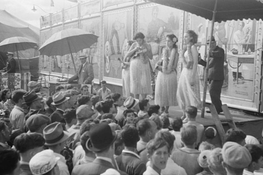 Black and white photo of a bustling carnival or sideshow scene from the early to mid-20th century. Three women in sparkling dresses stand on a platform under an awning, possibly performers or part of a “girlie show,” as a large crowd of mostly men in hats looks on. Behind them, colorful painted panels depict caricatured figures and sensational scenes, suggesting the presence of a “freak show” attraction, common during this era.