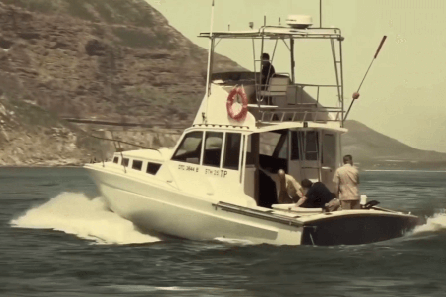 White motorboat named "STH 25 TP" speeds through the water with a visible wake, as several people onboard tend to someone or something at the stern. Rocky hills and coastline form the scenic background under clear skies.
