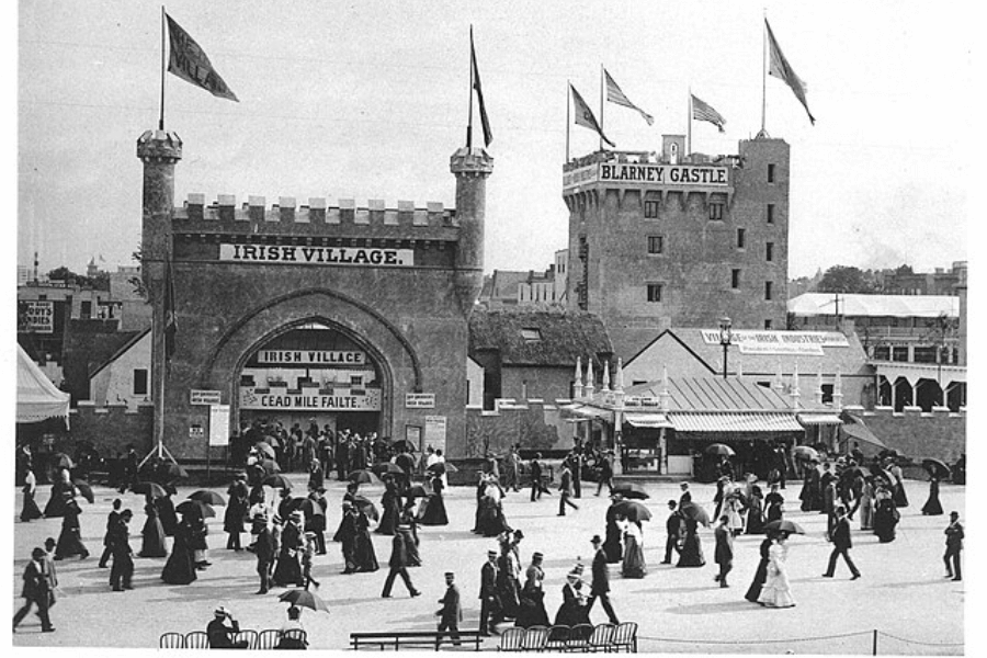 Black and white photograph of a grand entrance labeled "Irish Village" with castle-like towers and flags, part of a World's Fair or exposition. A large sign also marks “Blarney Castle” in the background. Crowds of men and women in Edwardian dress, many with parasols, stroll in front of the display, capturing a scene of cultural exhibition and early 20th-century public entertainment.