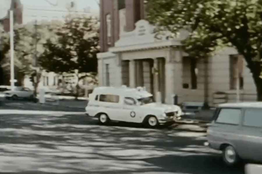 Vintage scene of a white ambulance-style vehicle with a red cross emblem driving through a quiet city street, with trees casting shadows and older buildings lining the background. Another light-colored car is seen passing in the foreground.