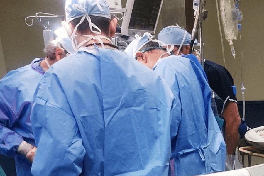A team of surgeons in blue scrubs and caps gathered around an operating table, focused on a medical procedure. Various medical monitors and IV lines are visible, indicating a fully equipped surgical environment.