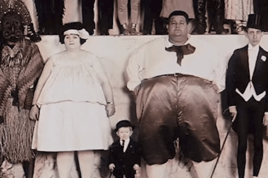 Colorized vintage photograph of a group of sideshow performers posed for a publicity photo. Among them are a man with extreme obesity, a woman labeled as a “fat lady,” a little person in formalwear, a man in a tribal costume and mask, and a tall thin man in a tuxedo. The image reflects the exploitative "freak show" era, when individuals with physical differences or non-Western heritage were exhibited for public amusement.