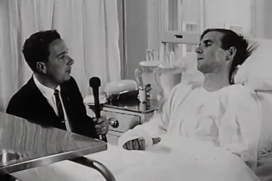 Black-and-white photo showing a suited man holding a microphone and interviewing a patient lying in a hospital bed. The patient appears to have bandaged hands and is propped up with pillows, suggesting a formal hospital setting.