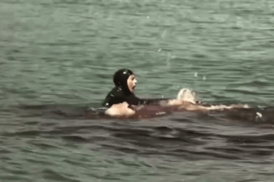 A diver in a black wetsuit screams in panic while holding onto a second person floating face-up in the water, surrounded by a large pool of red, likely blood. The scene conveys urgency, danger, and possible injury or attack.