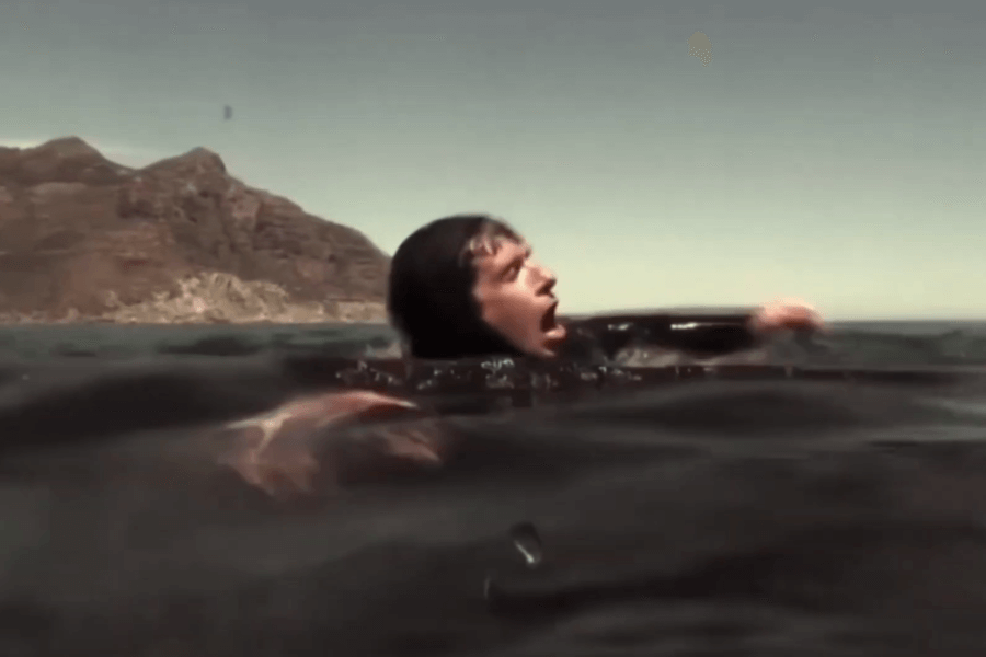 A person in a wetsuit floats in open water near a mountainous coastline, mouth wide open in a scream and arms raised in panic. The water around them is dark, hinting at a tense or dangerous situation.