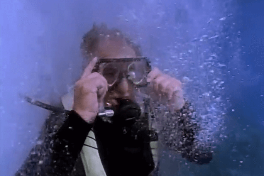 A scuba diver adjusts their mask underwater while surrounded by a cloud of bubbles, indicating active movement or air release. The diver is wearing a wetsuit and breathing apparatus in a deep blue marine environment.