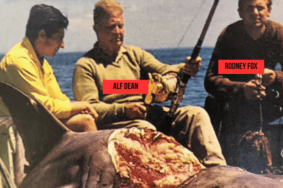 Color photo of three men on a boat with fishing rods, sitting behind a large, partially butchered shark. Labels identify Alf Dean in the center holding a golden reel and Rodney Fox on the right. The man on the left is unnamed, wearing a yellow shirt.