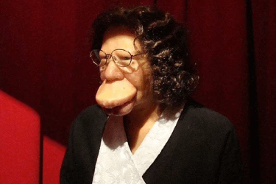 Photograph of a life-size wax figure modeled after Grace McDaniels, known for her severe facial disfigurement caused by Sturge-Weber syndrome. The figure wears glasses, a curly brown wig, and a black outfit with a white collar, posed against a deep red background. The exhibit likely aims to represent her historical presence in sideshow culture, though it underscores how such figures have often been displayed through a lens of spectacle rather than humanity.