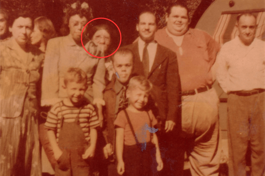 Colorized vintage group photo of sideshow performers and circus community members, gathered outdoors in front of a trailer. A red circle highlights Grace McDaniels, a woman with severe facial disfigurement from Sturge-Weber syndrome, standing among the group. The people around her include individuals with visible physical differences, such as dwarfism and obesity, reflecting the tight-knit yet exploited world of sideshow life in mid-20th century America.