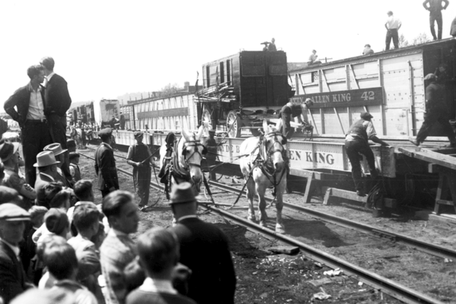 Black and white photo capturing a busy circus train unloading scene, with horses pulling ornate wagons down ramps from flatbed railcars labeled “Al G. Barnes” and “Allen King.” Workers and handlers guide the animals while crowds of onlookers, including many children, gather along the train tracks to watch. The moment reflects the behind-the-scenes labor of traveling circuses in the early 20th century and the public fascination with spectacle and mobility.