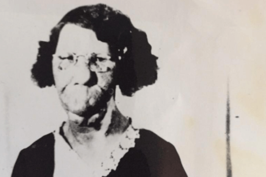 Faded black and white portrait of Grace McDaniels, a woman with a pronounced facial disfigurement caused by Sturge-Weber syndrome. She is wearing round glasses, a dark dress with a lace-trimmed collar, and looks calmly at the camera. The image’s simplicity and stark contrast emphasize her strength and visibility, offering a powerful reminder of her humanity amid an era of exploitation.