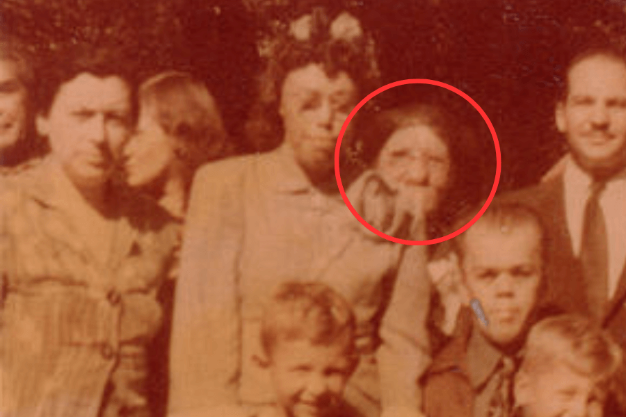 Vintage color photograph of a group of people posing together, with a red circle highlighting a woman near the center who has significant facial deformities. Her condition, possibly related to a genetic disorder or untreated illness, stands in stark contrast to the rest of the smiling group—underscoring historical neglect and the visibility of difference within mainstream society.