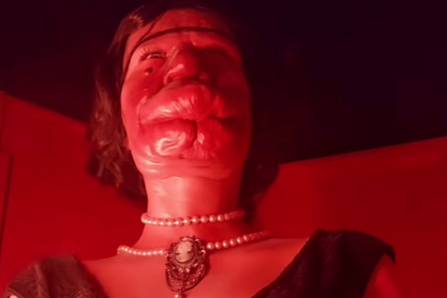 Low-angle photo of a wax figure illuminated in intense red light, modeled to depict Grace McDaniels, a woman with severe facial disfigurement caused by Sturge-Weber syndrome. The figure wears a pearl necklace with a cameo pendant and a dark dress. The lighting casts sharp shadows and creates an eerie atmosphere, evoking the sensationalized framing often used in sideshow exhibits, while prompting reflection on the ethics of visual representation.