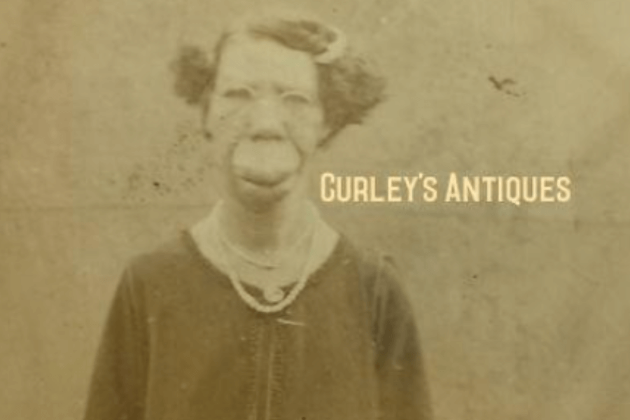 Sepia-toned portrait of Grace McDaniels, a woman with a prominent facial disfigurement caused by Sturge-Weber syndrome. She wears a dark dress with a light collar and a string of pearls, standing against a plain studio backdrop. The watermark “Curley’s Antiques” appears across the image, indicating it is part of a vintage or resale archive. The portrait captures both her visibility and dignity amid a history of public exhibition.