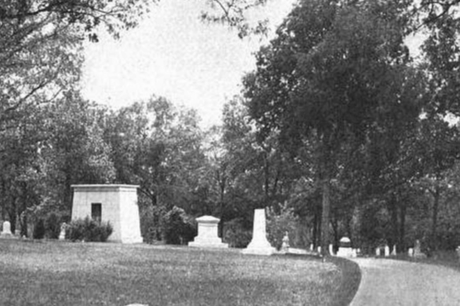 Black and white photograph of a peaceful cemetery with a winding path bordered by large trees and various monuments, obelisks, and tombstones. The headstones range in size and style, with some large mausoleum-like structures in the foreground. The tranquil setting evokes a sense of reverence and remembrance, appropriate for honoring the lives of those once marginalized or misunderstood.