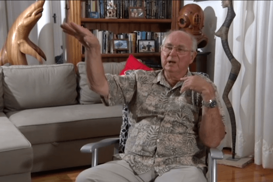 An elderly man wearing glasses and a patterned shirt sits in a living room, gesturing with one hand raised and the other touching his chest, as if explaining something. The background features bookshelves, a model diving helmet, ocean-themed decor, and a wooden dolphin sculpture.