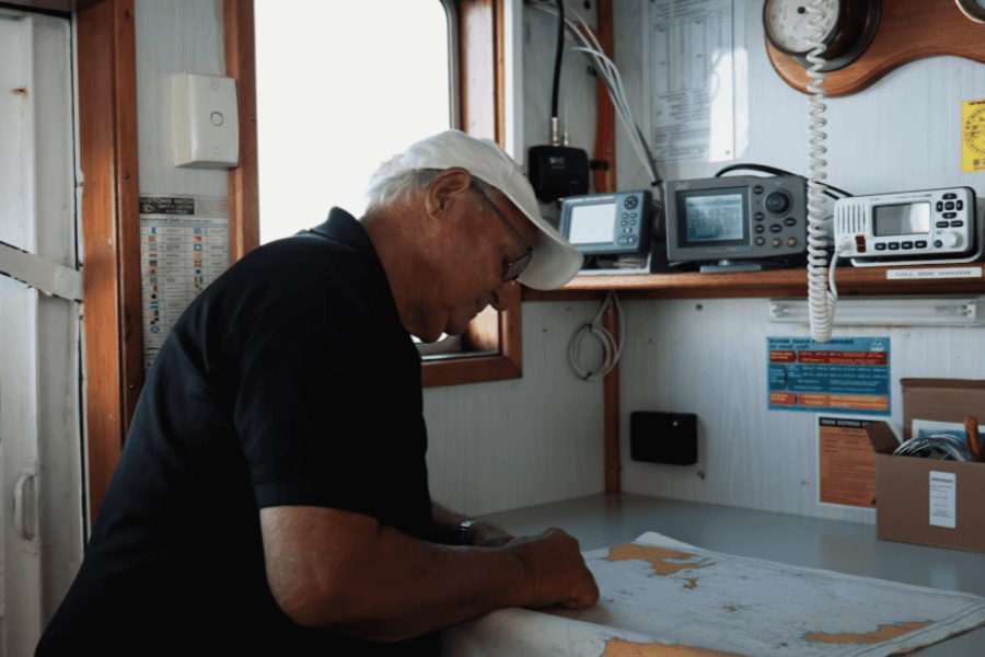 An elderly man wearing a white cap and black polo shirt studies a nautical chart inside a boat’s cabin. The space is equipped with marine radios, navigation instruments, and safety notices pinned to the wall.