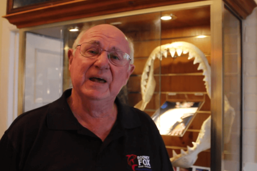 An older man wearing a black Rodney Fox-branded shirt stands in front of a glass display case containing a large set of shark jaws. The man appears to be mid-sentence, likely explaining or presenting something related to shark conservation or history.