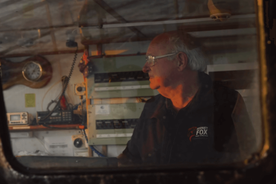 An older man in a "Rodney Fox Shark Expeditions" jacket stands inside a boat's control room, viewed through a window. Surrounded by navigation instruments and communication gear, he gazes out thoughtfully, illuminated by warm, ambient light.