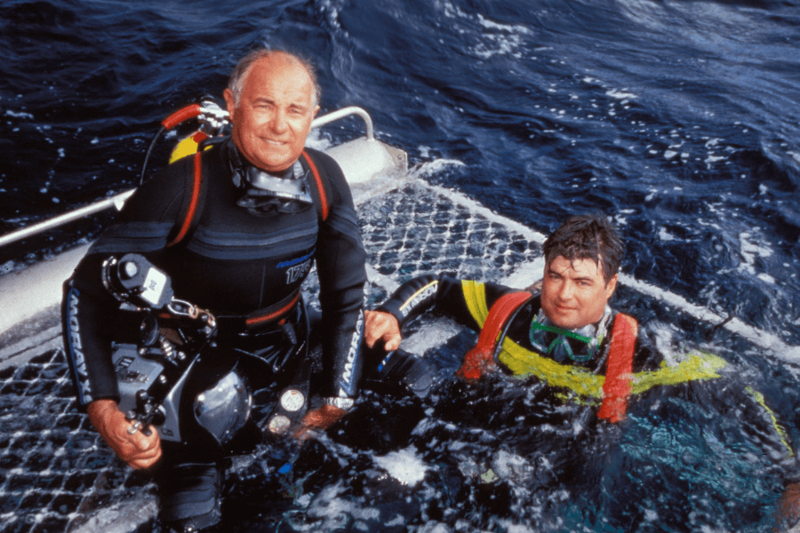 Two divers in full wetsuits prepare for or emerge from a dive next to a shark cage platform in the open ocean. One diver, holding an underwater camera rig, is seated on the platform, while the other floats in the water wearing a snorkel mask and red harness.