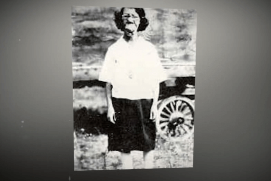 Black and white photo of a woman standing outdoors, wearing a white blouse and dark skirt, with a noticeable facial deformity that suggests a possible congenital or medical condition. Behind her is a wagon wheel, evoking a rural or early 20th-century setting, emphasizing the historical marginalization of individuals with visible differences.