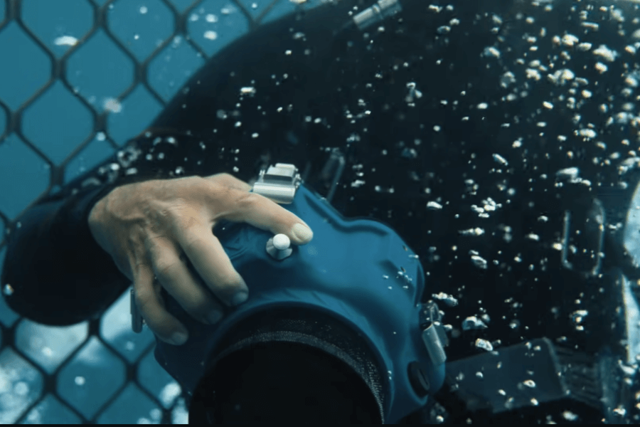 Close-up of a diver’s hand operating a professional underwater camera encased in a blue waterproof housing. The diver is submerged, surrounded by air bubbles, with a shark cage grid visible in the background.