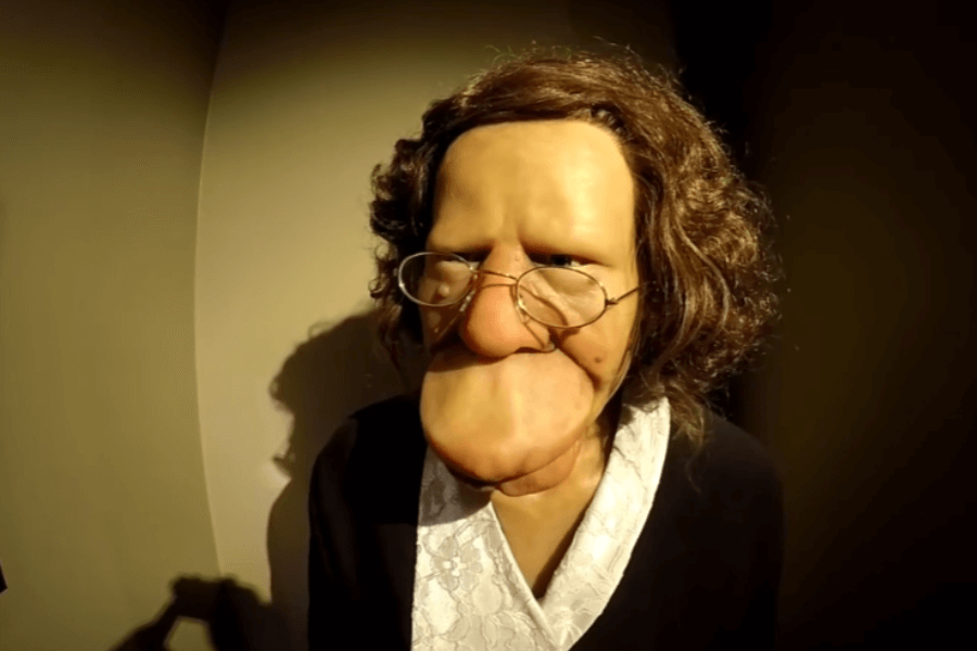 Photograph of a wax figure modeled after Grace McDaniels, a woman who lived with severe facial disfigurement caused by Sturge-Weber syndrome. The figure wears a black dress with a white lace collar and round glasses, with curly brown hair. The soft lighting and neutral background contrast with the figure’s exaggerated facial features, offering a reflection on how disability was once displayed for spectacle rather than dignity.