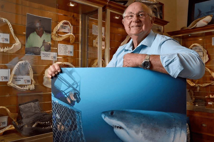 An older man, smiling and wearing glasses and a light blue shirt, stands in a shark museum surrounded by jaws on display, holding a large underwater photo of a great white shark near a diver in a cage. The man appears to be Rodney Fox, noted shark attack survivor and conservationist, featured in a poster and photo in the background.