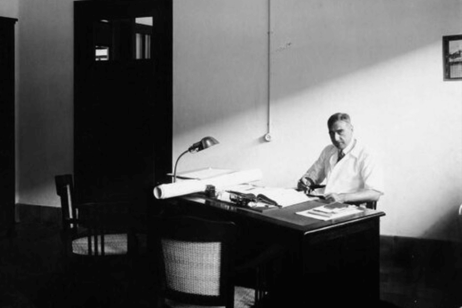 Black and white photo of a man seated at a wooden desk in a minimalist, well-lit office. He is wearing a white shirt and looking toward the camera, surrounded by architectural blueprints, books, and a desk lamp. A beam of sunlight cuts diagonally across the room, emphasizing the stark simplicity and contemplative mood of mid-20th century professional life.