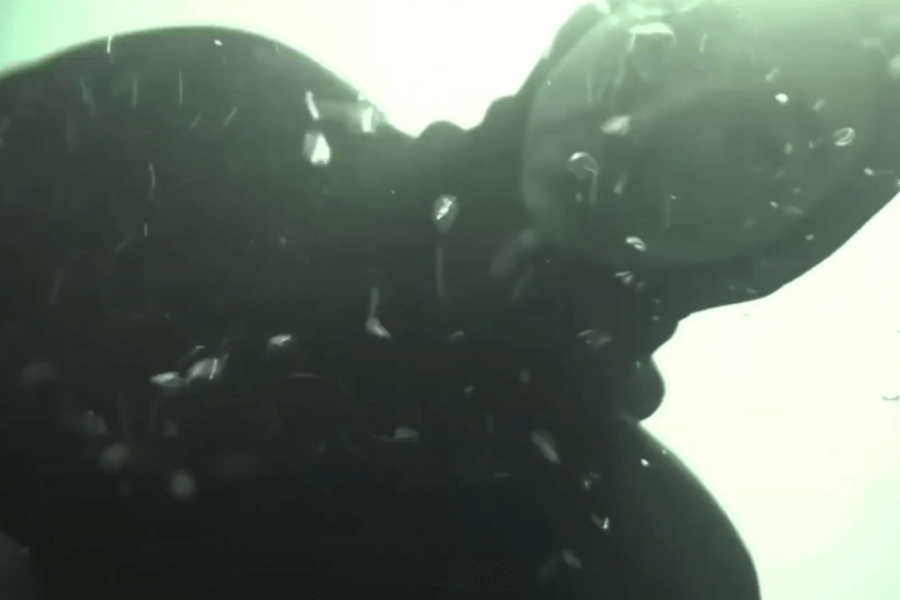 Underwater view of a diver in a wetsuit and full-face mask surrounded by rising air bubbles, with light filtering from the surface above. The diver’s face is partially visible through the mask, creating a moody and immersive atmosphere.