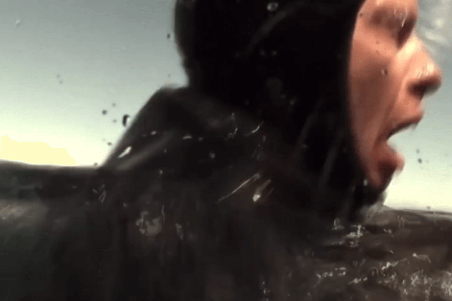 Close-up of a diver in a black wetsuit emerging from the water with a panicked expression, mouth open as if shouting or gasping. Water splashes around their shoulder, suggesting urgency or fear.