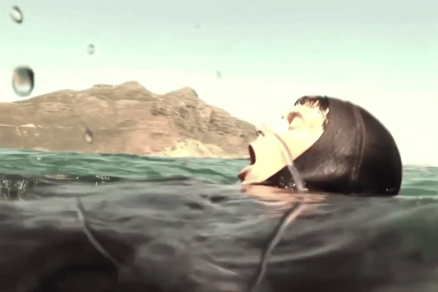 A diver in a black wetsuit floats in open water with their mouth open, appearing to gasp for air or call out. The rocky coastline and sunlit mountain are visible in the background under a clear sky.