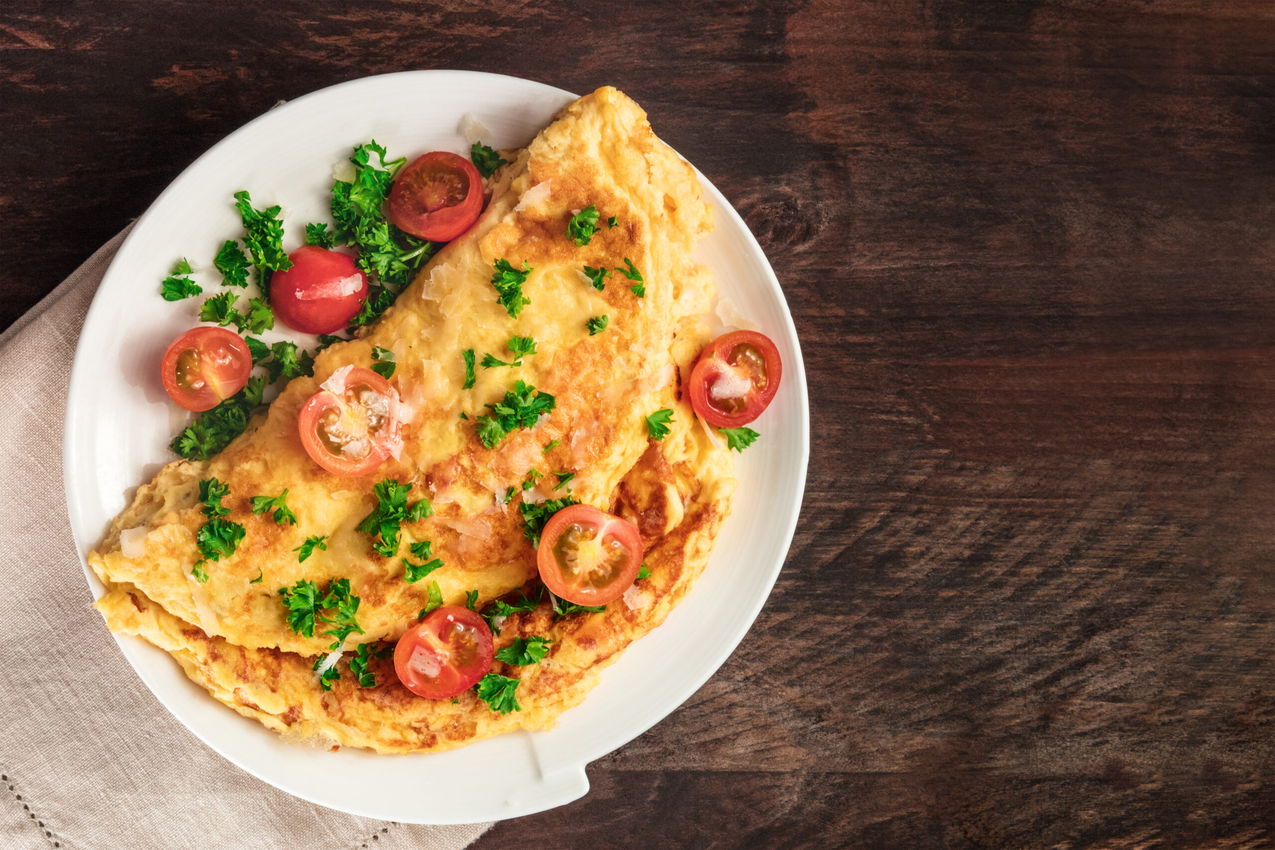 A photo of an omelet with cherry tomatoes, parsley. and grated cheese, shot from above on a rustic wooden texture with a place for text