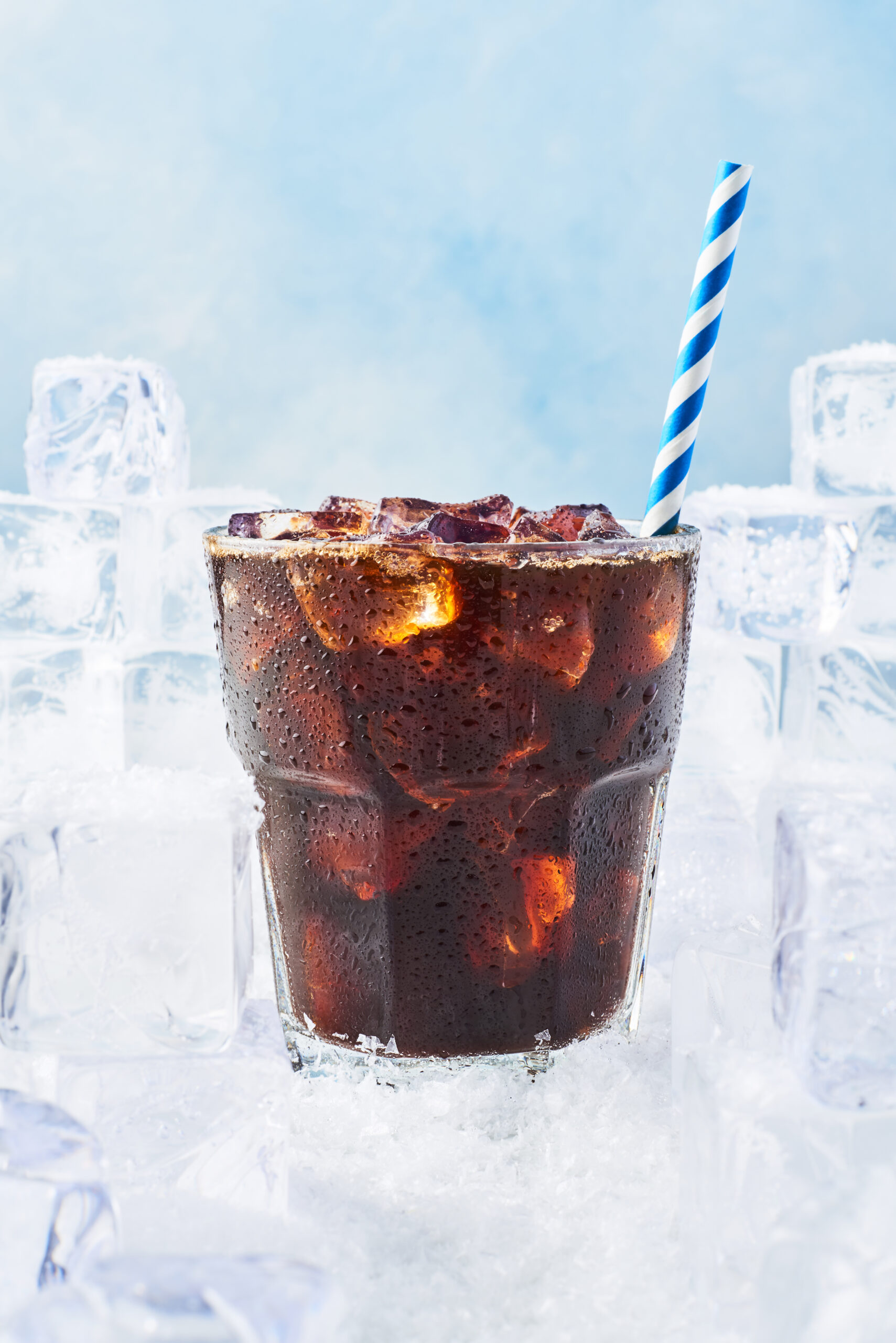 Summer drink iced coffee or soda in a glass with droplets and straw surrounded by ice cubes on snow over blue background. Selective focus, copy space for text.