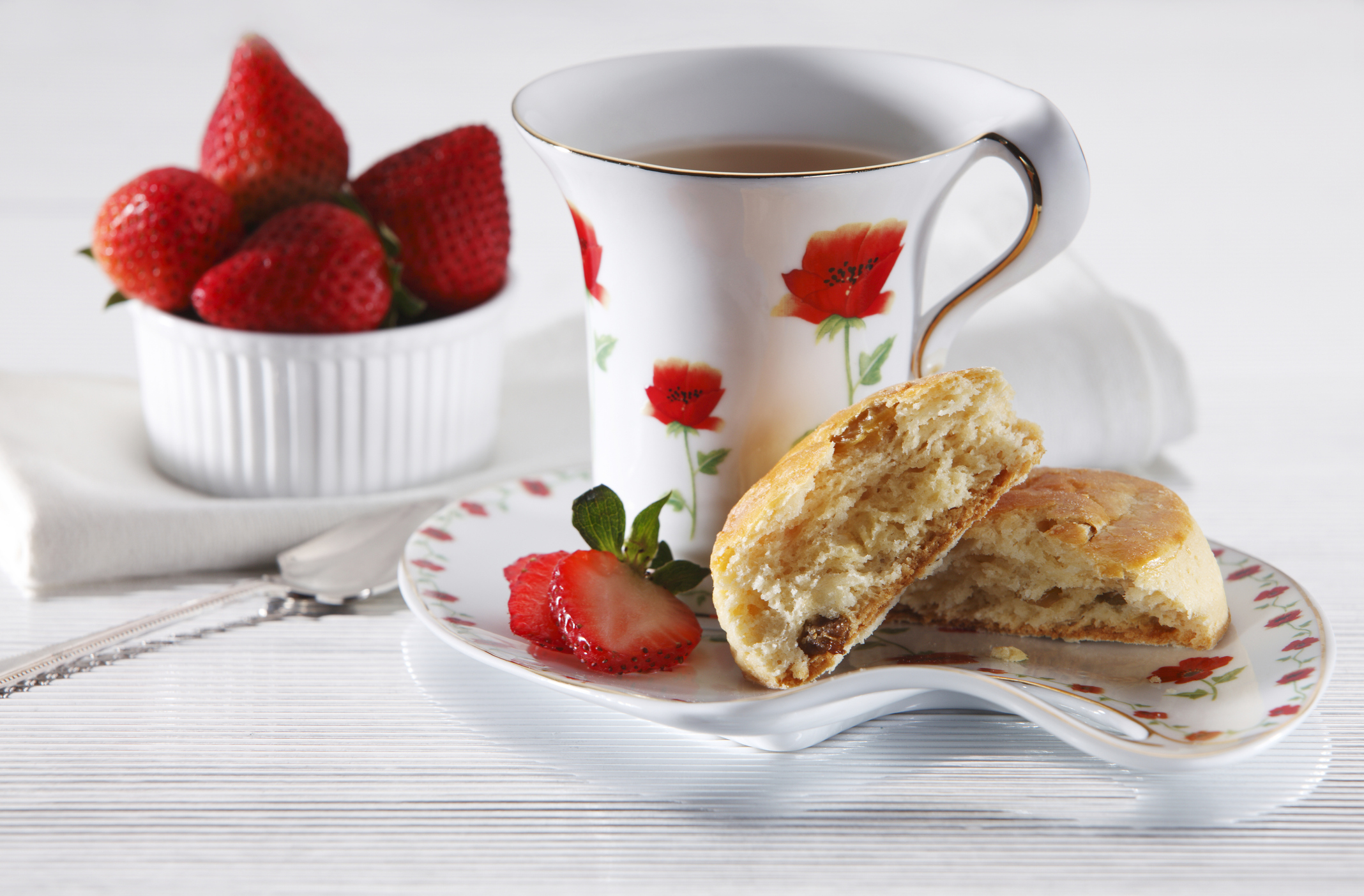 Fresh strawberries in a white dish next to a strawberry-themed teacup and a scone sitting on a white dish.