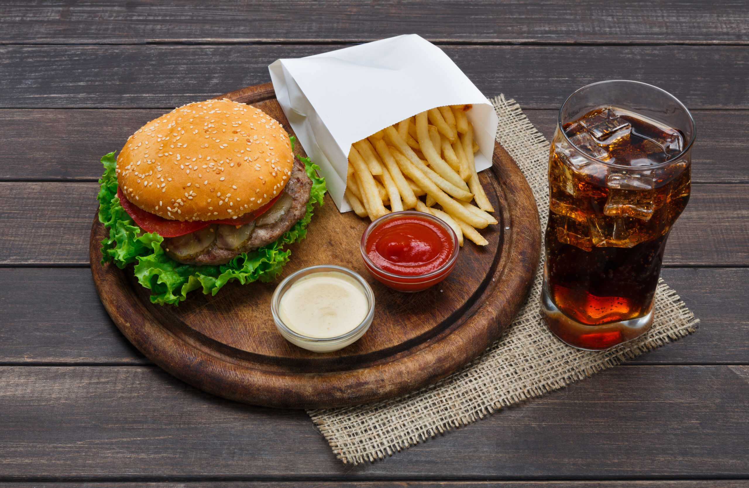 Fast food dish. Meat burger, potato chips and glass of cola drink with ice on wood. Takeaway composition. Wrapped French fries, hamburger, mayonnaise and ketchup sauces on wooden desk.