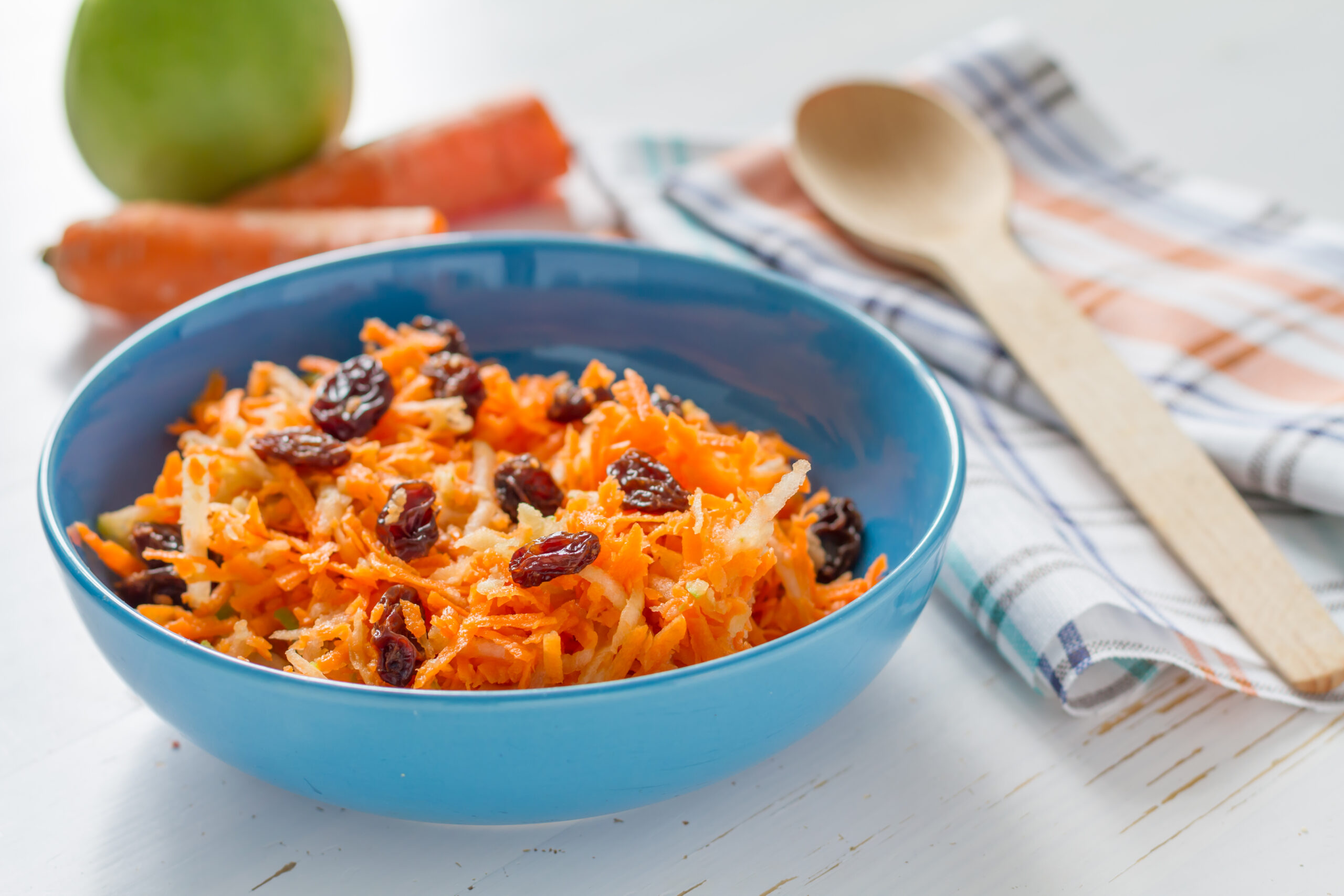 Salad - carrot, apple and raisins in blue bowl with wood spoon on white wood background