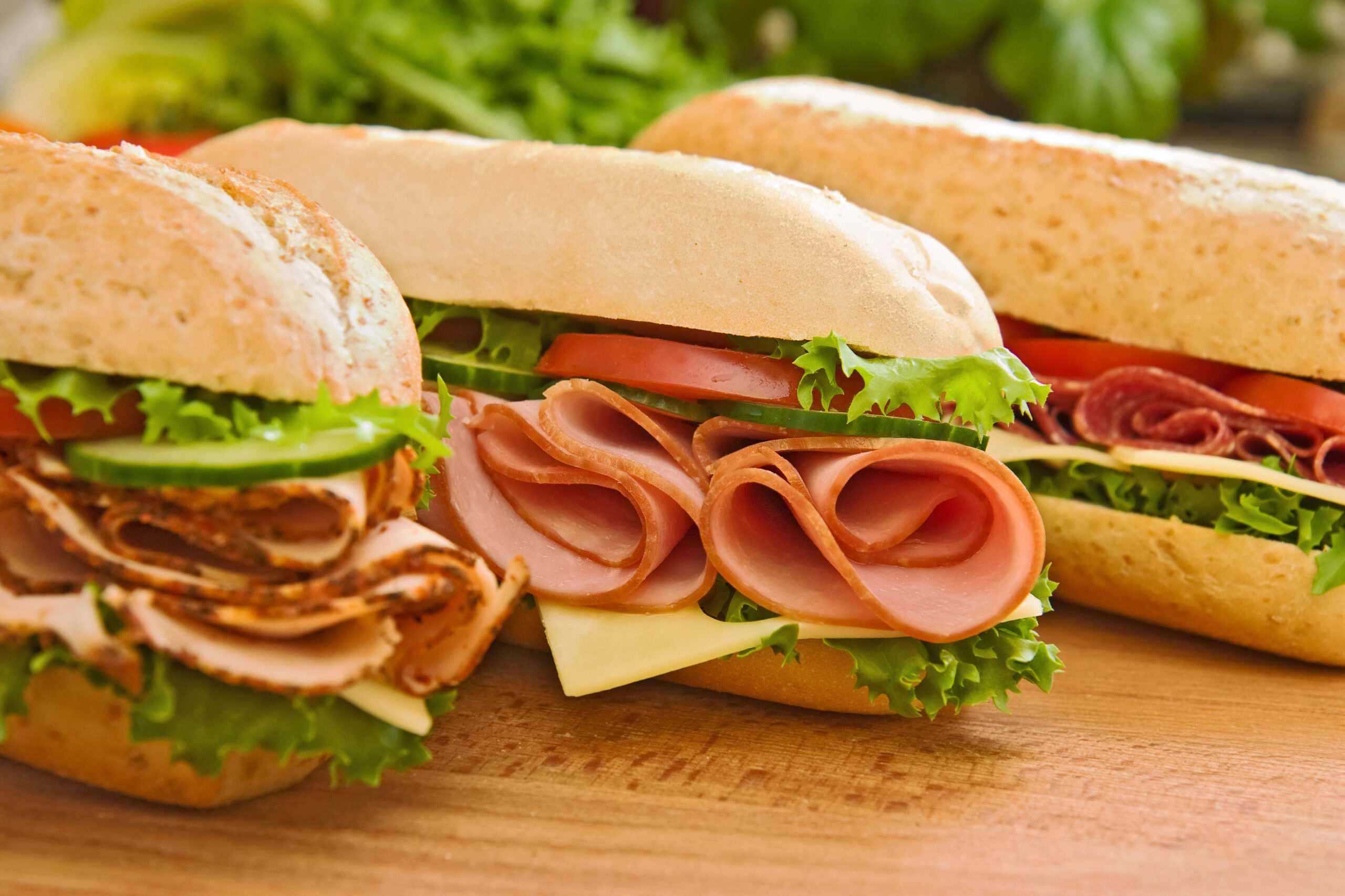Three fresh sub sandwiches - turkey/chicken breast, ham & swiss and salami on a cutting board. Focus on the ham sandwich