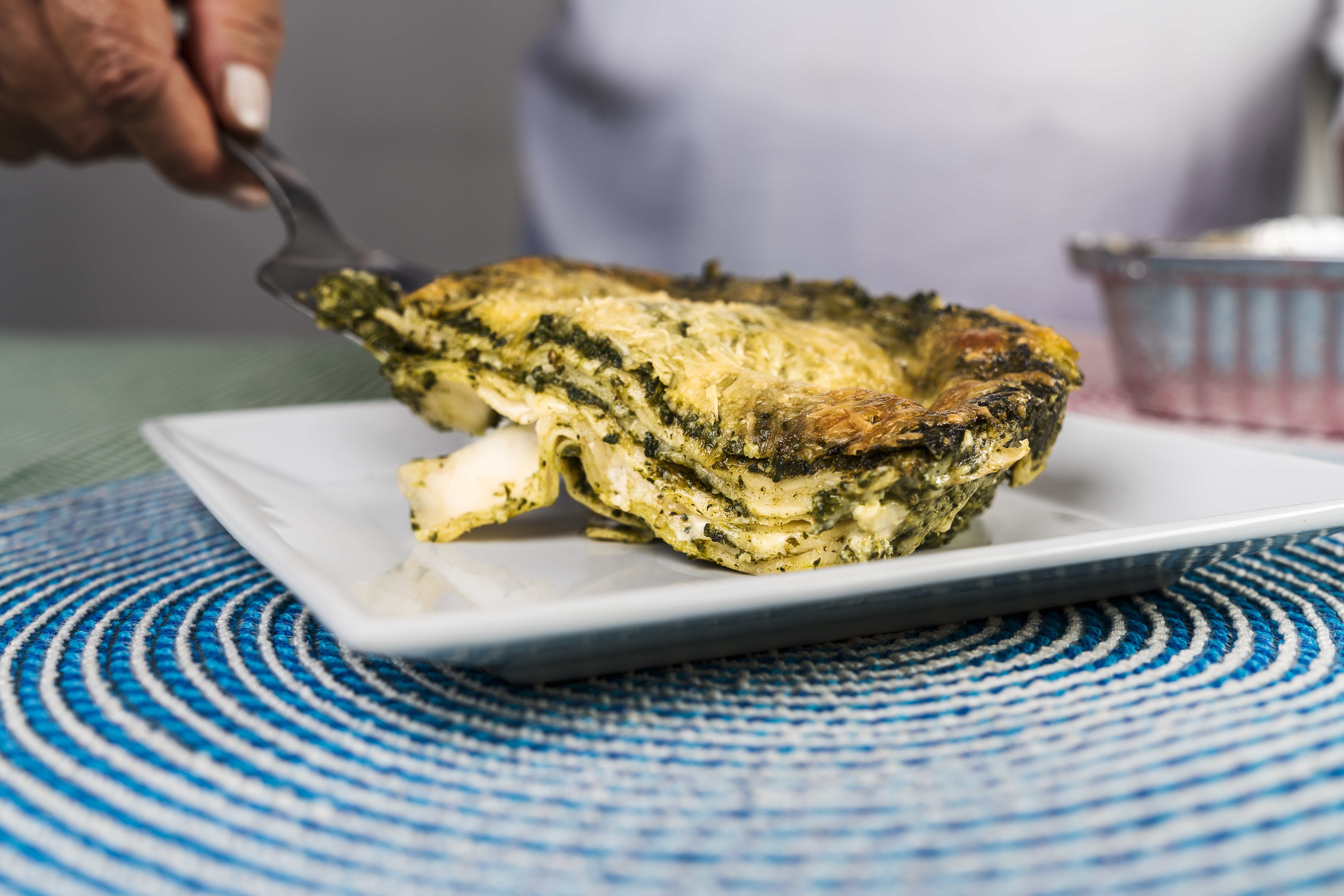 Close up view of a hand of an adult woman serving a portion of freshly made pesto lasagna on a plate.
