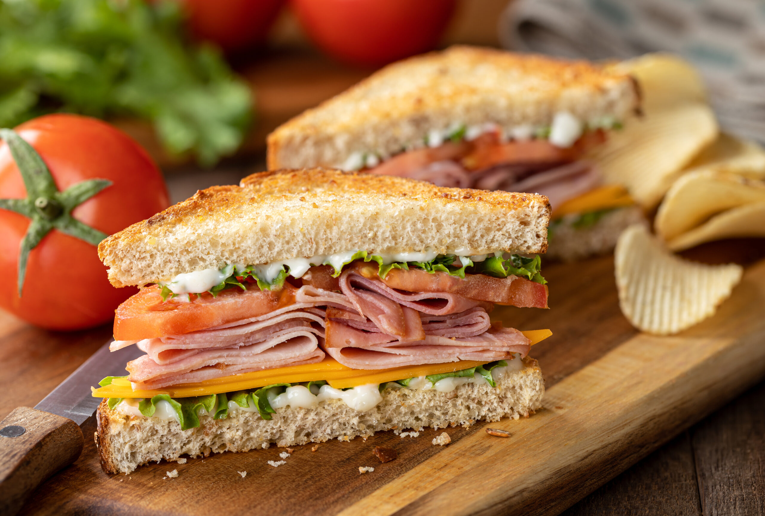 Ham sandwich with cheese, lettuce and tomato on whole grain bread.  Lettuce and tomatoes in background