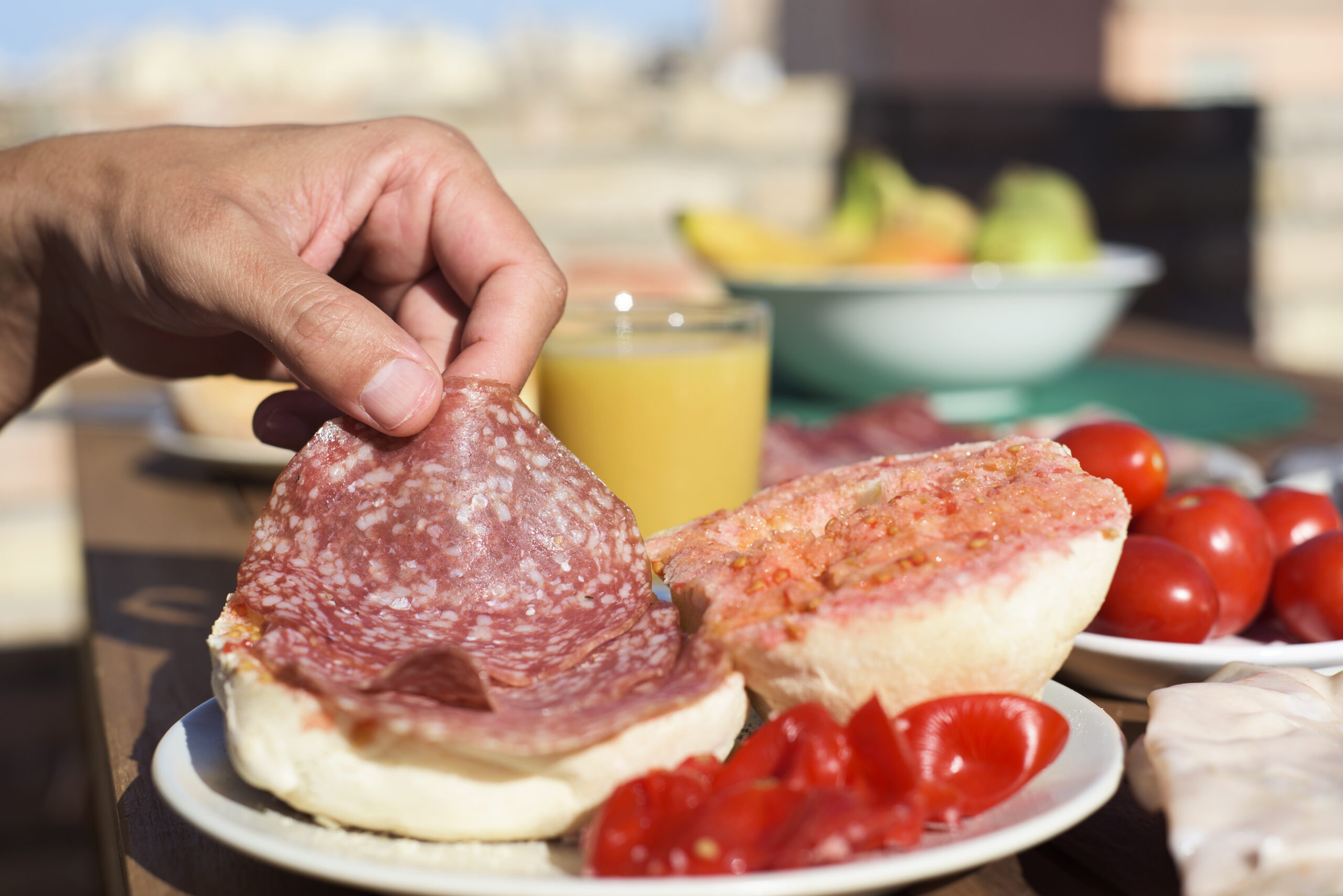 closeup of a man preparing a sandwich in a bun with typical catalan pa amb tomaquet, bread with tomato, stuffed with salami, on a wooden table outdoors