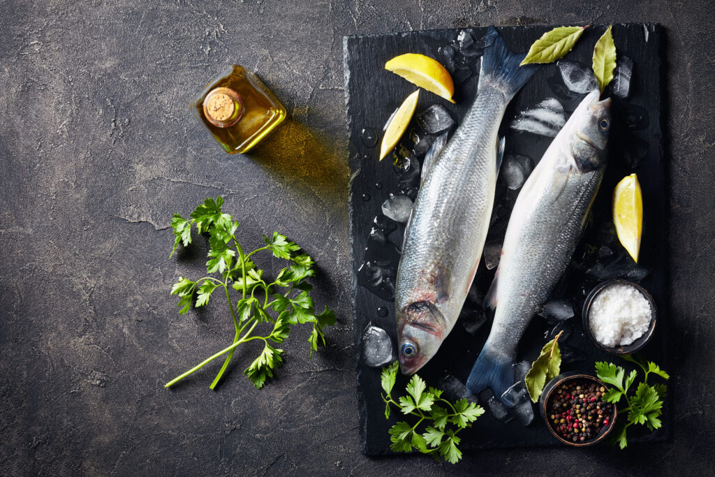 two fresh raw whole sea bass with lump ice, lemon slices, spices and herbs on a slate plate on a concrete table, view from above, close-up, flatlay, copy space