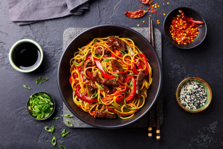 Stir fry noodles with vegetables and beef in black bowl. Slate background. Top view.