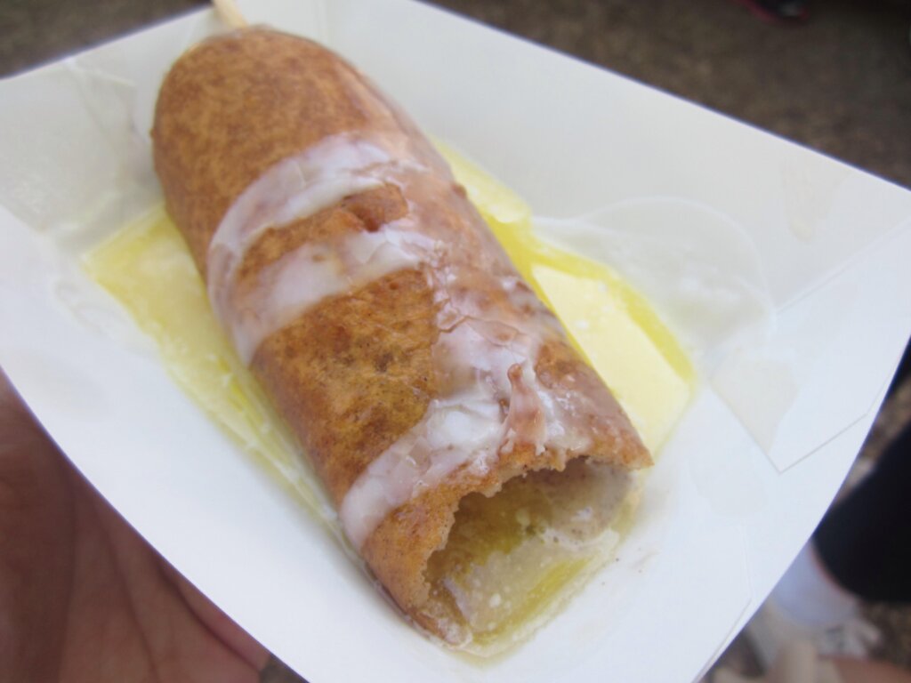 A deep-fried stick of butter coated in a golden-brown batter, drizzled with icing, and served on a tray with a pool of melted butter underneath. The batter is cracked open at the front, revealing the melted interior.