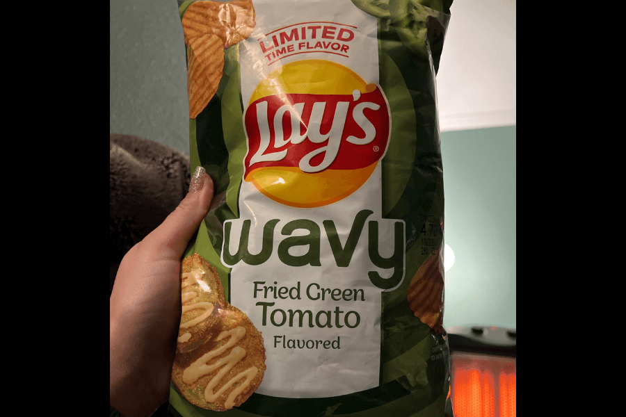 A bag of Lay's Wavy chips in the flavor "Fried Green Tomato," marked as a limited-time flavor. The packaging features green and white colors, with an image of fried green tomato slices topped with a drizzle of sauce. A hand is holding the bag against a softly lit indoor background.