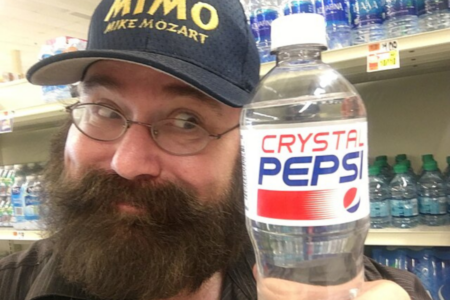 This image shows a person in a store holding a bottle of Crystal Pepsi in front of shelves stocked with bottled water. The individual has a beard, wears glasses, and is dressed in a "MIMO Mike Mozart" cap, smiling slightly while showcasing the product.