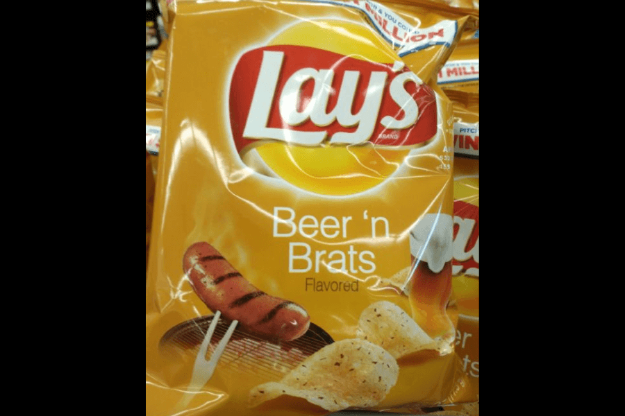 A bag of Lay's chips in the "Beer 'n Brats" flavor, featuring a golden-yellow design. The packaging displays an image of a grilled bratwurst on a fork alongside seasoned potato chips, with a frothy glass of beer in the background. The bag is part of a store display with other Lay's bags visible behind it.